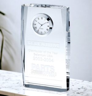 A crystal Plaque Clock that's made with a thick crystal plaque with beveled edges. It features a 2" clock with a white face & silver bezel. The bottom area of the clock has laser engraving personalization with a company logo & words for the distributor of the year. This crystal clock is 4" x 7" in size & weighs 3.4 lbs. It's sitting on a white marble shelf.