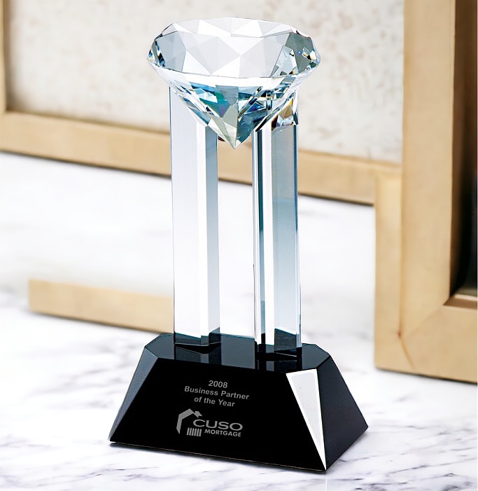 Our Venus Diamond Award that features a 4" solid crystal diamond resting on 2 crystal pillars. The pillars are mounted on a black crystal base which includes a black & silver engraving plate for personalization. This crystal award is sitting on a white marble shelf.