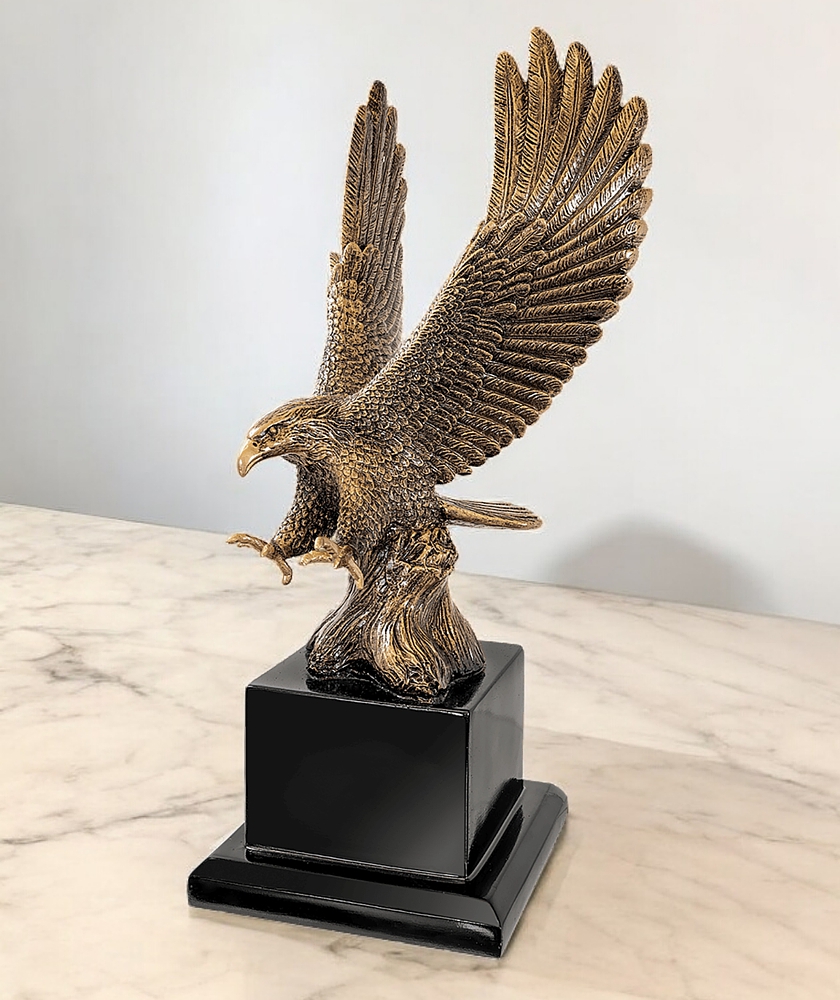 Our Eagle In Flight Statue features an antique gold bald eagle with his wings spread & talons out ready to attack. It's mounted on a black wood base with that includes an engraving plate for personalization. (Not shown here). This EGL80AG eagle statue is turned to the left to give you a side profile look. It's sitting on a cream marble table.