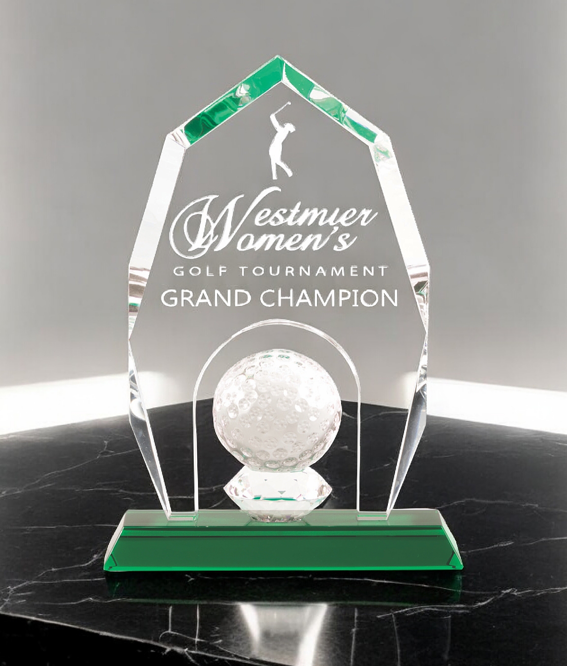 Our Crystal Apex Golf Trophy CRY955. It features a clear piece of crystal with an apex peak at the top and a solid crystal golf ball at the bottom for decoration. The middle area is for laser engraving personalization and the base is made from green crystal. It stands 9.5" tall & weighs 4 lbs. This trophy is sitting on a black marble table with a grey wall in the background.