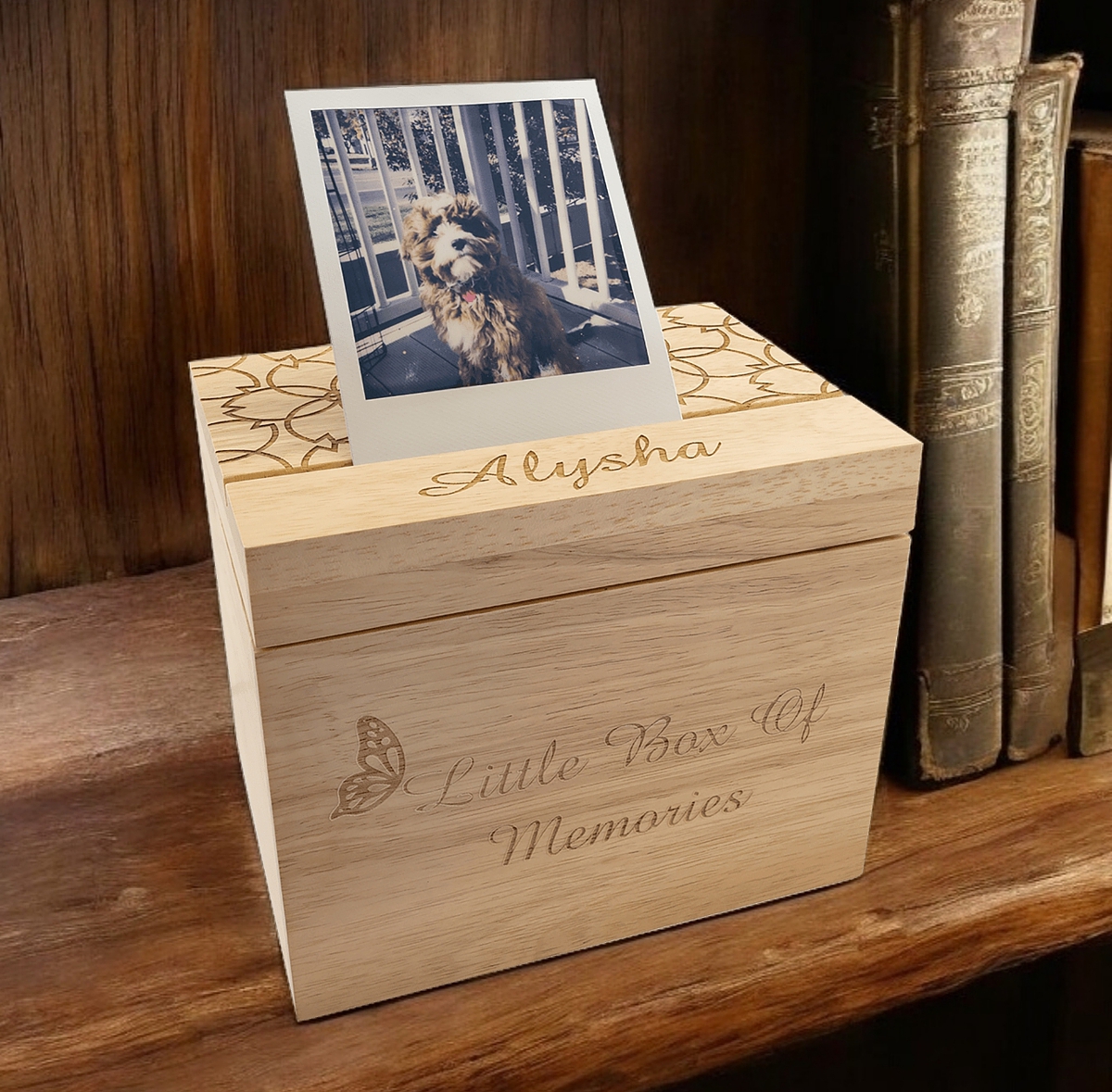 Here's a photo of our Personalized Keepsake Box being used as a dog memorial box. It's sitting on a shelf with books to the right. The box is engraved with the dog's name Alysha and a polaroid photo of the dog is standing in a slit in the lid of the box.