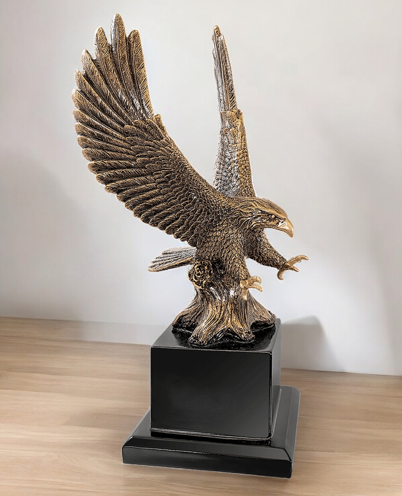 Our Eagle in Flight Statue turned to the left so you can get a different side profile look. It's sitting on a light wood table with a white wall in the background. This EGL80AG eagle sculpture is made with resin and has an antique gold look to it. It's mounted on a black wood base that includes an engraving plate that can be personalized, but one is not shown in this photo.