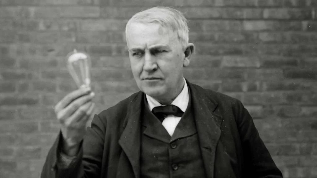 A black & white photo of Thomas Edison holding a light bulb and looking at it intensely. 