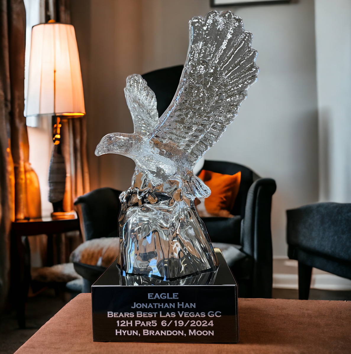 Our Crystal Eagle Statue features a clear crystal eagle with his eagles spread wide mounted on a black base. The base includes a black & silver engraving plate with personalization on it. This K9117 eagle statue is sitting on a wood desk in front of a chair & lamp.