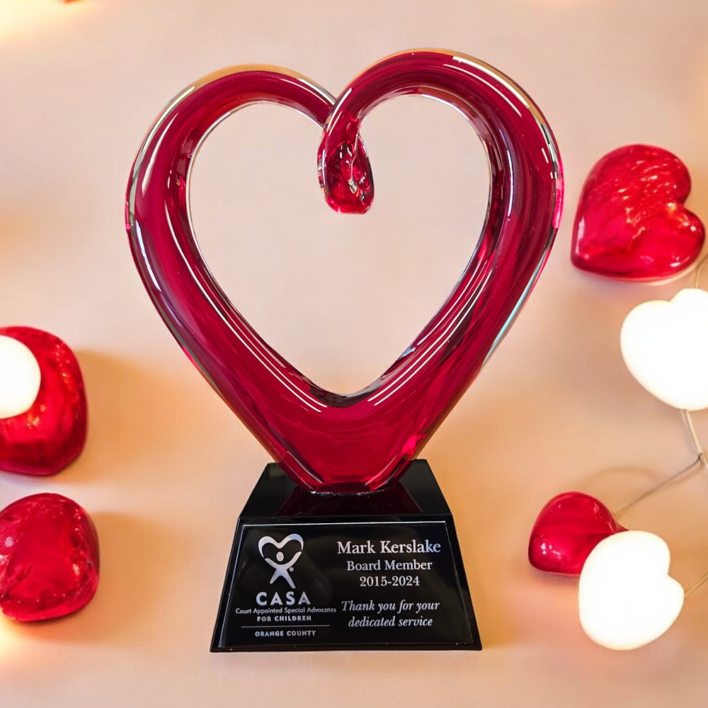 The Whole Heart Art Glass Award features a red glass heart made from blown glass. It's mounted on a black glass base with a black & silver engraving for personalization. It stands 9" tall, weighs over 5 lbs & comes in a satin-lined deluxe gift box. This specific art glass piece is sittting on a table with white & red heart lights for decoration.