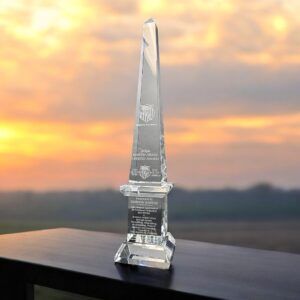 Our Large Crystal Obelisk Award sitting on a black table outside with a sunset in the background. This CRY121 crystal award stands 24" tall and weighs 21 lbs. This specific award is personalized for Gordon Santee for winning the AAU USA Martin Drake Legend Award