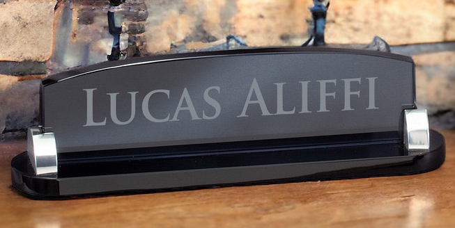 Our Smoked Glass Desk Name Plate laser engraved with a name, sitting on a desk with a light brick background.