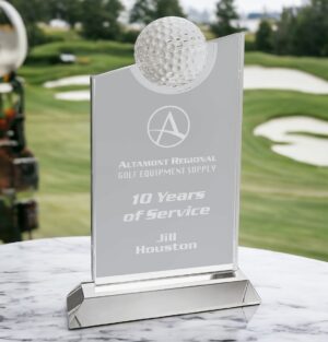 Our Slanted Golf Ball Trophy is made with a clear crystal rectangle with a slanted top, going downward from right to left. Also in the middle of that slant is a 2.5" crystal golf ball for added decoration. Below that is a blank area for laser engraving personalization. This one is engraved for someone who has been at a golf company for 10 years. The golf trophy is sitting on a white marble table and the background shows a golf course with some sand traps.