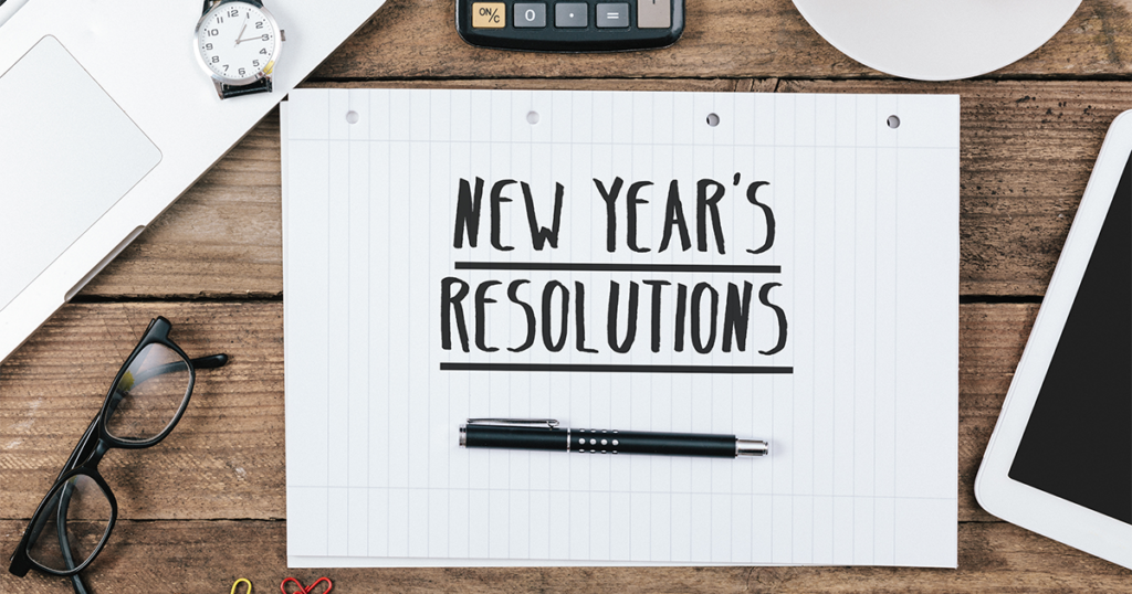 A tablet of paper reading, "New Years Resolutions" with a pen below. The tablet is on a wooden desk with items such as glasses, clock, calculator & tablet around it. 