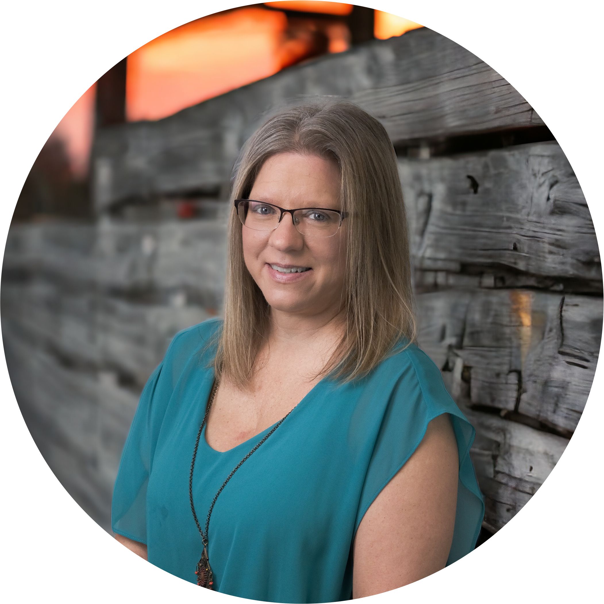 Brooke Snow is an employee of Hit Trophy. Her job is to laser engrave trophies & awards. She's shown here in a teal top in front of a rustic wood fence and a sunset in the background.