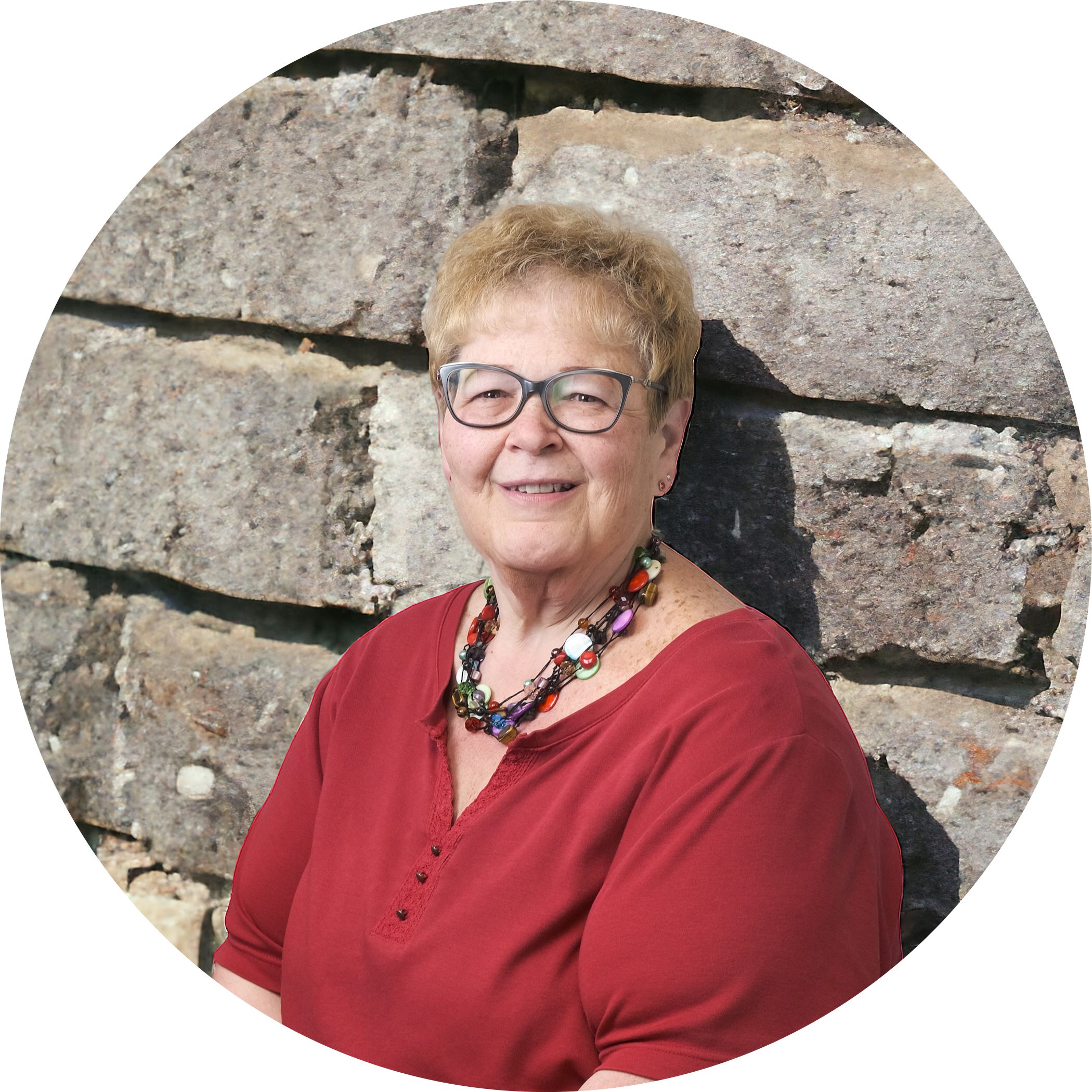 Kelly Schmucker, the office administrator of Hit Trophy in a red blouse, colorful necklace & retro glasses. She's leaning up against a decorative block wall.