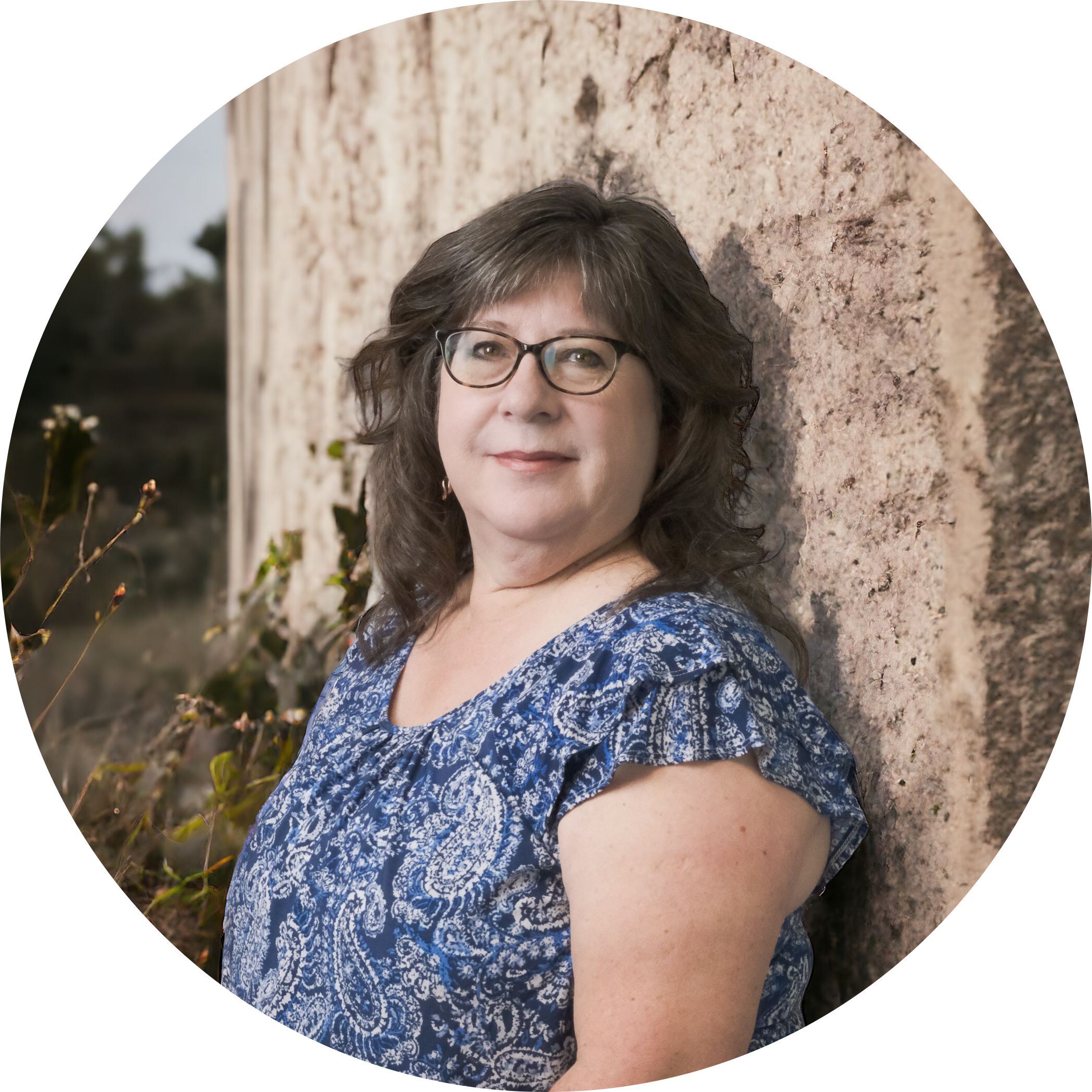 Valerie Kinsman is an employee of Hit Trophy. Here she is in a blue flower blouse as she's leaning up against a decorative concrete wall with some plants and a field in the background.