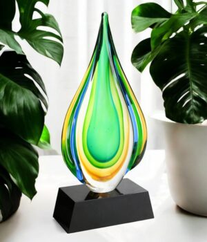 Our Rainforest Art Glass Award sitting on a white table with tropical plants on both sides. The award features a raindrop shaped piece of glass with rainforest themed colors throughout. It's mounted on a black glass base that includes an engraving plate for personalization. This G1808B is 13" tall & weighs 6.2 lbs.